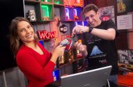 Mulher de camisa rosa posa em escritório. Monitores de computador visíveis no fundo.