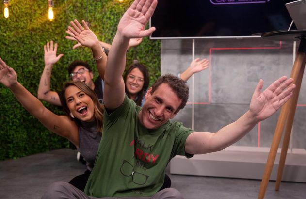 Dois homens sorrindo em estúdio. Um segura dado branco, e outro dado na mesa.