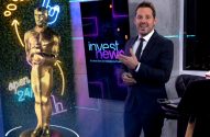 Homem de terno apresenta Invest News em estúdio com estátua dourada e luzes neon.