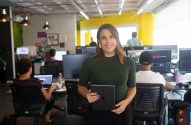 Mulher sorrindo segura tablet em escritório movimentado com pessoas trabalhando em computadores.