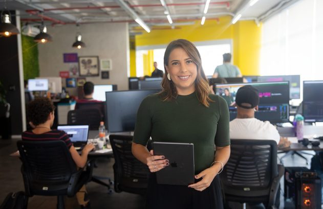 Mulher sorrindo segura tablet em escritório movimentado com pessoas trabalhando em computadores.