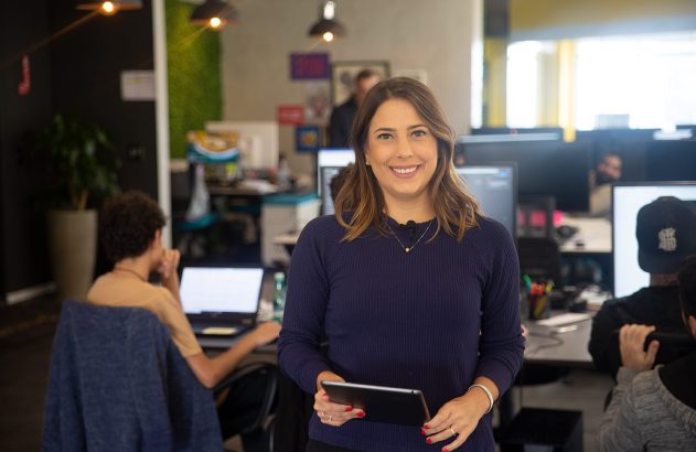 Mulher sorrindo, segurando tablet, em escritório com colegas trabalhando.