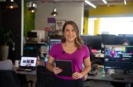 Mulher sorridente com microfone e tablet em um escritório, com sua imagem em um monitor.