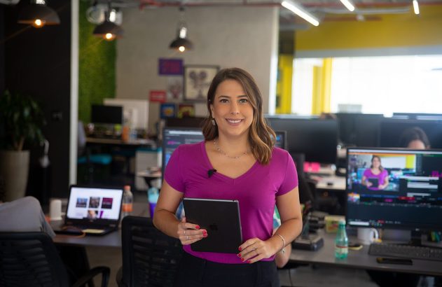 Mulher sorridente com microfone e tablet em um escritório, com sua imagem em um monitor.