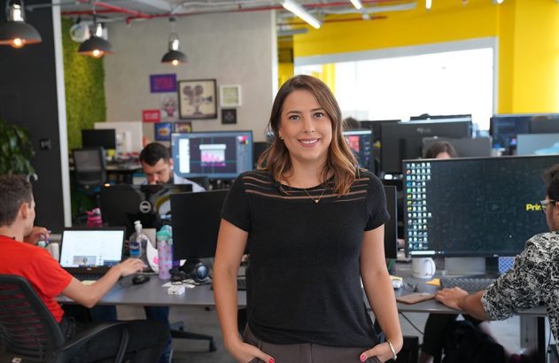 Mulher sorridente em escritório movimentado com pessoas trabalhando em computadores.
