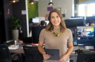 Profissional sorridente em escritório moderno, segurando tablet e olhando para a câmera.
