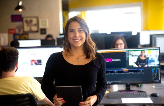 Mulher sorridente segurando um tablet em escritório com telas de computador ao fundo.