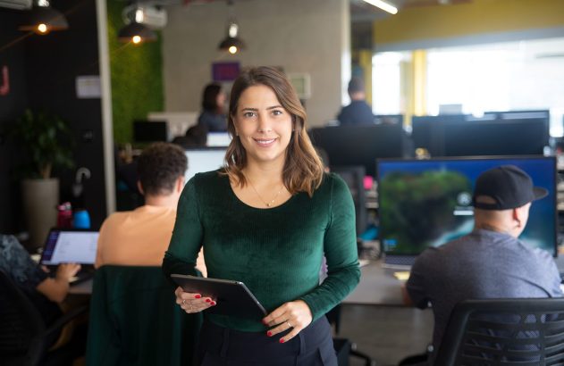 Mulher sorrindo com um tablet em um escritório com pessoas trabalhando.