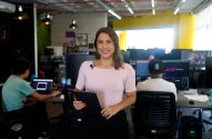 Mulher sorridente em escritório, com funcionários trabalhando ao fundo.