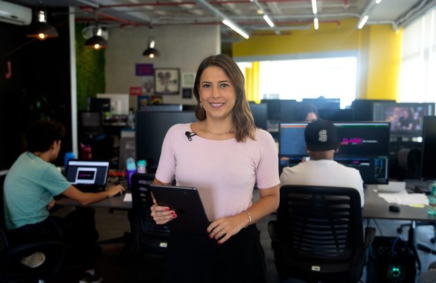 Mulher sorridente em escritório, com funcionários trabalhando ao fundo.