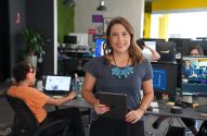 Mulher sorridente segura tablet em escritório. Um monitor exibe sua imagem; pessoas trabalham em PCs ao fundo.