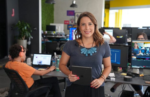 Mulher sorridente segura tablet em escritório. Um monitor exibe sua imagem; pessoas trabalham em PCs ao fundo.