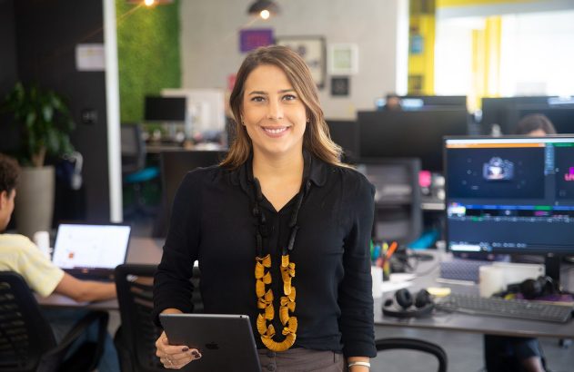 Mulher sorridente em escritório, segurando tablet. Pessoas e computadores ao fundo.