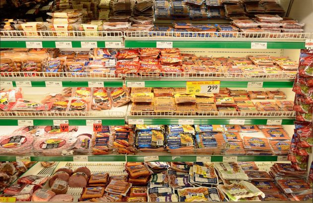 Expositor de supermercado com salsichas e frios embalados em prateleiras refrigeradas, com preços visíveis.