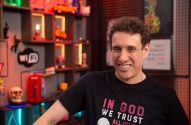 Homem de cabelo cacheado e camiseta preta sorrindo para a câmera, em estúdio com estante de itens decorativos.
