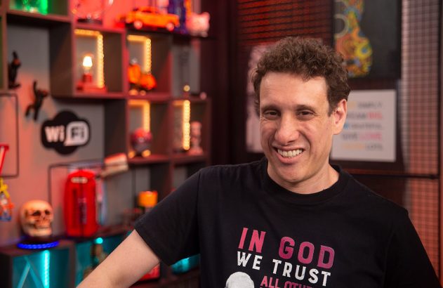 Homem de cabelo cacheado e camiseta preta sorrindo para a câmera, em estúdio com estante de itens decorativos.