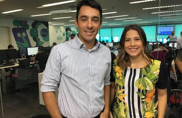 Homem e mulher sorrindo, posando em pé em uma redação. Ele de camisa, ela blusa colorida.
