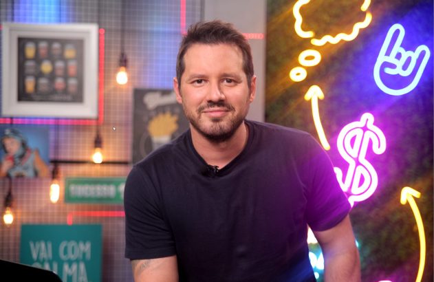 Homem sorridente com barba e camiseta preta posa em estúdio. No fundo, luzes neon de balão de fala, mão de rock e cifrão.