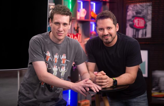 Dois homens sorrindo para a câmera, apoiados em mesa. Um veste camiseta cinza com gráficos, o outro preta.