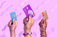 Três mãos erguem cartões coloridos com designs variados (estrela, personagem, corações) em fundo rosa.