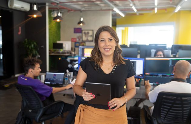 Mulher sorridente com tablet no centro de um escritório, com equipe trabalhando ao fundo.