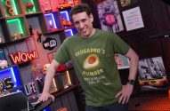 Homem sorridente em camiseta verde "Avogadro's Number" em fundo com luzes neon.