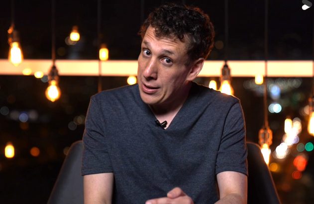 Homem sentado, olhando para a câmera com expressão falante, em fundo escuro com luzes.