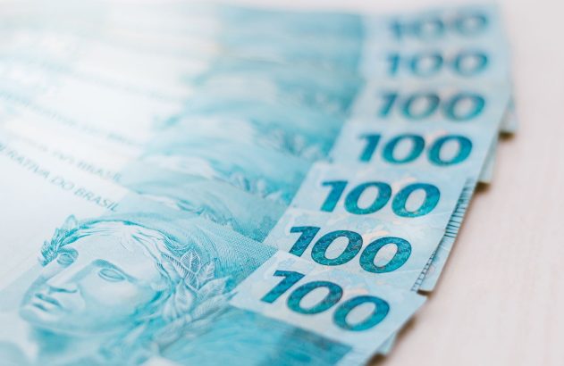 Cédulas de 100 Reais brasileiras em tons de azul e verde, parcialmente sobrepostas.