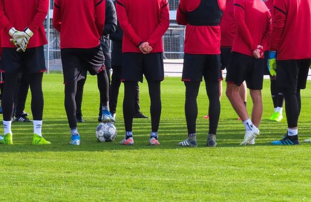 Jogadores de uniforme vermelho e preto de pé em campo de grama.