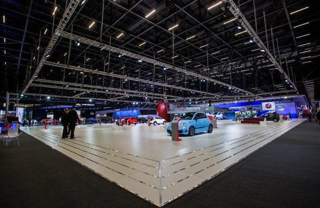 Exposição de carros com Fiat 500 azul em primeiro plano, pessoas e estandes de marcas como Fiat, Ford e Chevrolet.