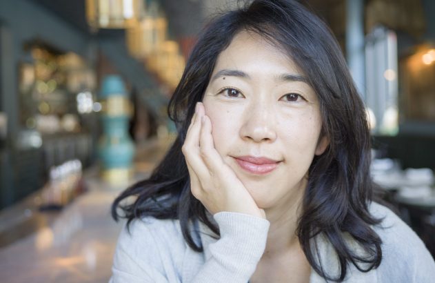 Mulher sorrindo, com a mão no queixo, em ambiente interno desfocado.