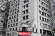 Letreiro digital "IMPOSTÔMETRO" em prédio branco de fachada curva, com edifício cinza ao lado.