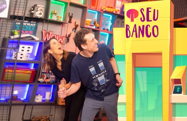 Mulher e homem surpresos, ela segurando o braço dele, olham para cima. Ao lado, um banco cenográfico.