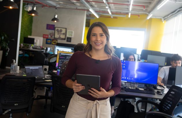 Mulher sorrindo segura um tablet em um escritório moderno com computadores ao fundo.