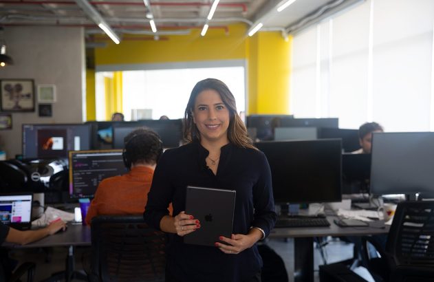 Mulher sorrindo segura tablet em escritório, com colegas trabalhando ao fundo.