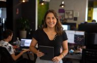 Mulher sorrindo segura tablet em escritório, com pessoas trabalhando ao fundo.