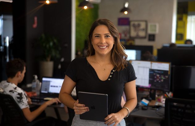 Mulher sorrindo segura tablet em escritório, com pessoas trabalhando ao fundo.