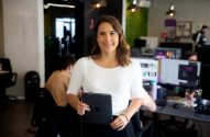 Mulher sorrindo, segurando um tablet, em um escritório com outras pessoas trabalhando.