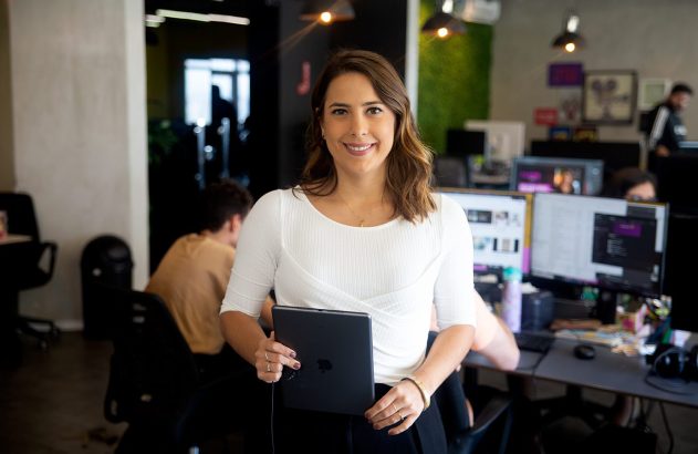 Mulher sorrindo, segurando um tablet, em um escritório com outras pessoas trabalhando.