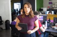 Mulher em pé, segurando pasta, em escritório com pessoas e computadores ao fundo.