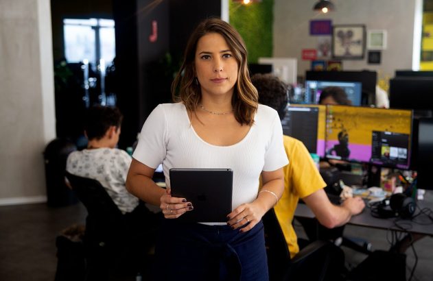 Mulher de camisa branca em pé no escritório, segurando tablet e olhando para a câmera.