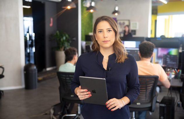 Mulher com tablet, olhando para a câmera, em ambiente de escritório com pessoas ao fundo.