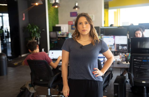 Mulher séria de camiseta azul-acinzentada, mão na cintura, em escritório com pessoas trabalhando ao fundo.