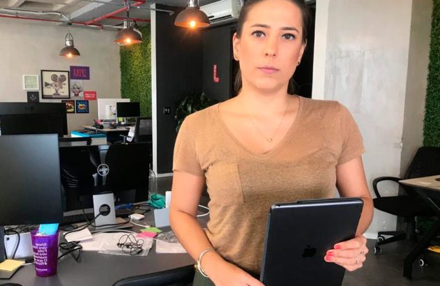 Mulher em pé, de camisa marrom, segurando um iPad em um escritório.