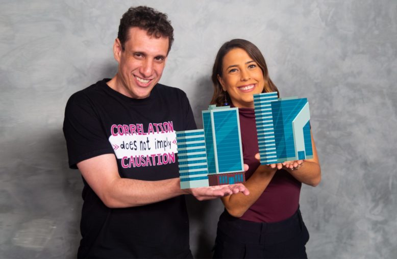 Um homem e uma mulher sorrindo seguram maquetes de prédios contra um fundo cinza.