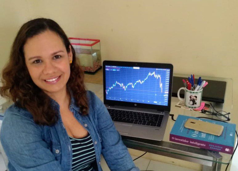 Mulher sorrindo em sua mesa, com gráfico financeiro exibido na tela de seu notebook.