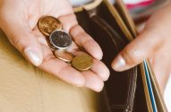 Mãos segurando moedas brasileiras sobre uma carteira aberta.