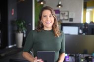 Mulher sorridente com blusa verde escura em escritório, segurando um tablet horizontalmente.