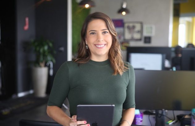 Mulher sorridente com blusa verde escura em escritório, segurando um tablet horizontalmente.