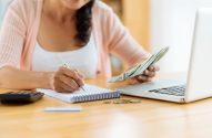 Pessoa gerenciando finanças: anota em caderno, segura dinheiro, com laptop, calculadora e moedas na mesa.